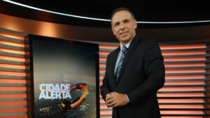 Reinaldo Gottino sorrindo com terno escuro e gravata ao lado de um monitor vertical exibindo o logo do Cidade Alerta na Record