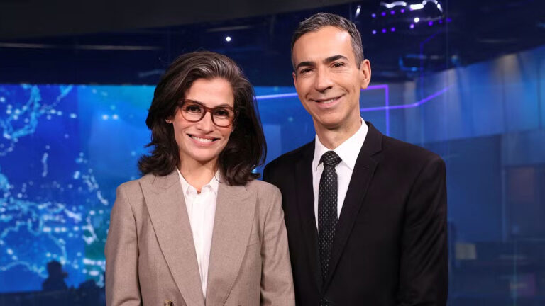 Renata Vasconcellos, de blazer bege, e César Tralli, de terno escuro, sorriem no estúdio do Jornal Nacional em frente ao telão azul