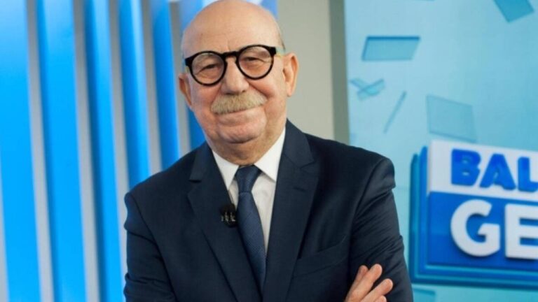 Homem sorridente de terno e gravata posa em estúdio de TV, com fundo azul e logotipo de programa jornalístico parcialmente visível