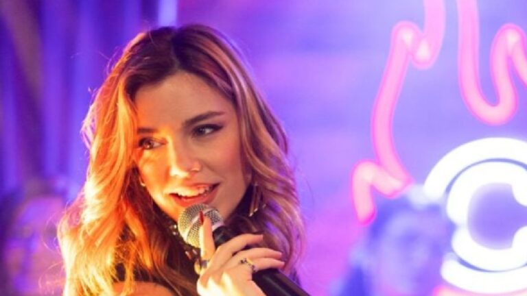 Mulher com cabelos soltos e maquiagem marcante segura um microfone e sorri enquanto canta em um palco iluminado por luzes coloridas e néon. O cenário sugere um ambiente de show ou apresentação musical noturna