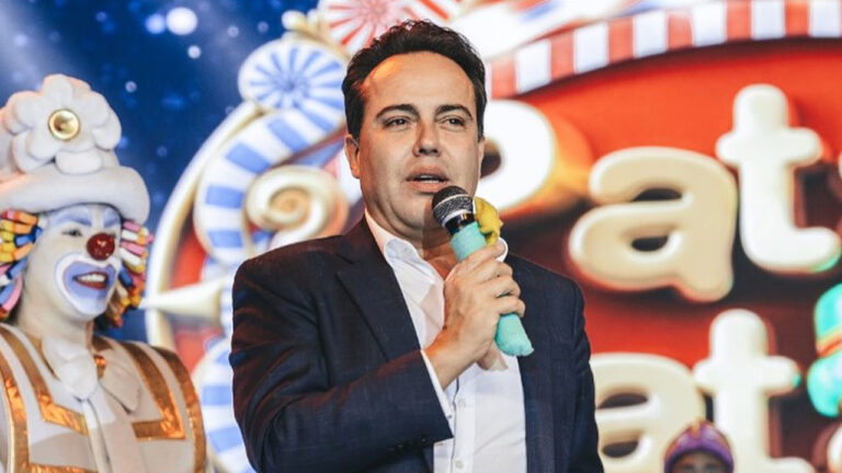 Rinaldi Faria, criador do Patati Patatá, falando ao microfone em um evento com fundo temático de doces e um palhaço