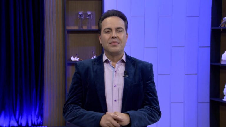 Rinaldi Faria, de blazer azul e camisa clara, aparece sorridente em estúdio de televisão iluminado por luzes azuladas. Ao fundo, há prateleiras com objetos decorativos e cortinas escuras, compondo um cenário elegante de programa de TV