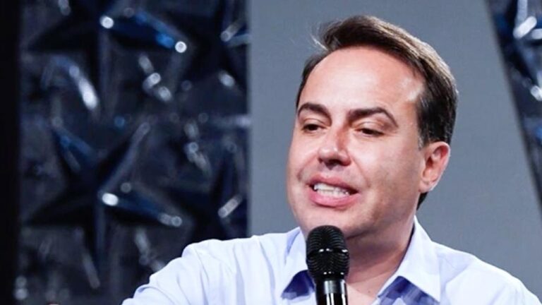 Homem de camisa social azul clara fala segurando um microfone em um ambiente interno, com painel decorado com estrelas ao fundo, sugerindo participação em um evento, palestra ou programa de TV