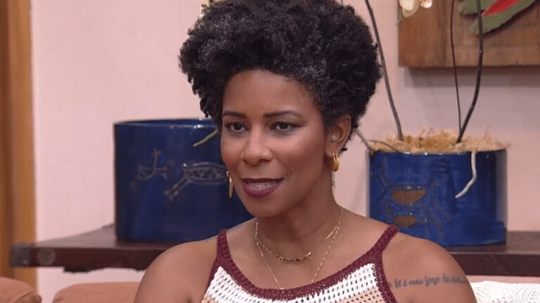 A imagem mostra uma mulher sentada em um ambiente interno, com expressão serena. Ela tem cabelo curto e cacheado, usa brincos dourados, colares delicados e uma blusa de crochê branca com borda vinho