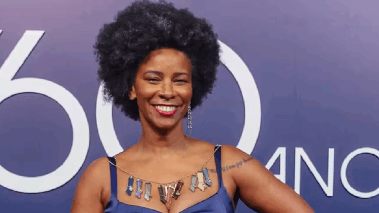 Rita Batista sorridente posa em frente a um painel roxo, vestindo um vestido azul e colar colorido, com cabelo em estilo afro e batom vermelho