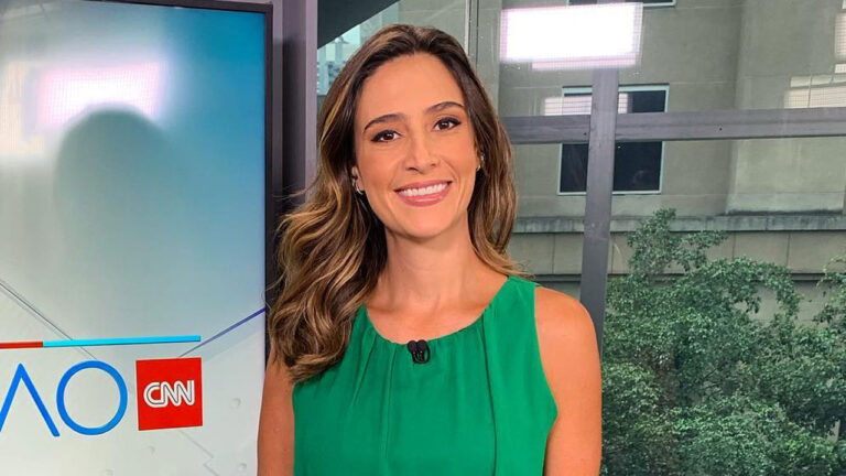 Jornalista Roberta Russo sorrindo, vestindo regata verde em estúdio da CNN Brasil com uma janela ao fundo da bancada