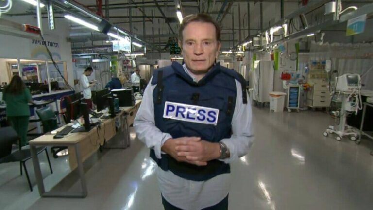 Roberto Cabrini usando colete azul com a palavra “PRESS” em destaque está em um ambiente hospitalar, com profissionais de saúde e equipamentos médicos ao fundo