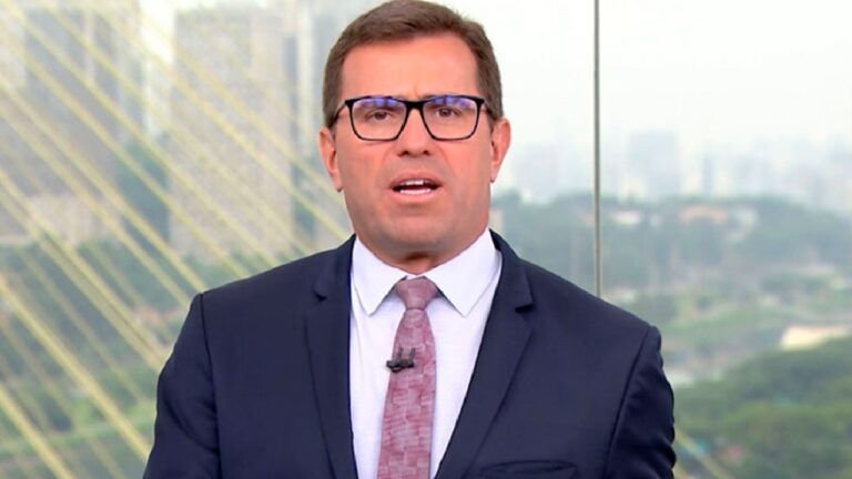 Homem de terno escuro, camisa branca e gravata rosa aparece falando em frente a um fundo urbano desfocado, com prédios e cabos de ponte visíveis. A imagem sugere a cena de um telejornal ou transmissão ao vivo de notícias