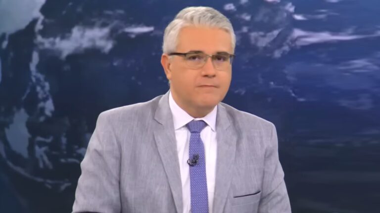 A imagem mostra Rodrigo Piscitelli, jornalista, no estúdio do Jornal da Cultura. Ele usa terno cinza-claro, camisa branca e gravata azul, e olha diretamente para a câmera com expressão séria. Ao fundo, há uma tela grande exibindo uma imagem do globo terrestre em tons de azul, típica de cenários jornalísticos