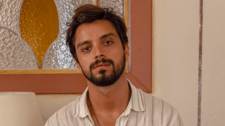 Ator Rodrigo Simas de barba e cabelo escuro, usando camisa listrada clara, posa em frente a uma janela de vidro