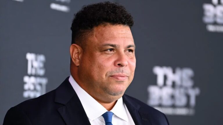 Homem de terno escuro e gravata azul posa em frente a painel do evento The Best