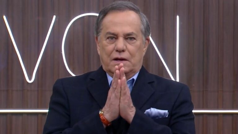A imagem mostra um homem de cabelo grisalho, vestindo um terno escuro e camisa azul clara. Ele está com as mãos unidas em frente ao rosto, em um gesto que pode indicar concentração ou ênfase no que está dizendo. Ao fundo, há um painel de madeira com letras iluminadas, sugerindo que ele está em um estúdio de televisão