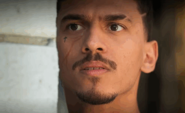 A imagem mostra um homem de expressão séria, encostado em uma parede clara. Ele tem um pequeno bigode e cavanhaque, além de uma tatuagem em forma de cruz próxima ao olho e uma cicatriz que desce pelo lado esquerdo do rosto. A iluminação destaca seu olhar atento e concentrado, sugerindo tensão ou expectativa na cena