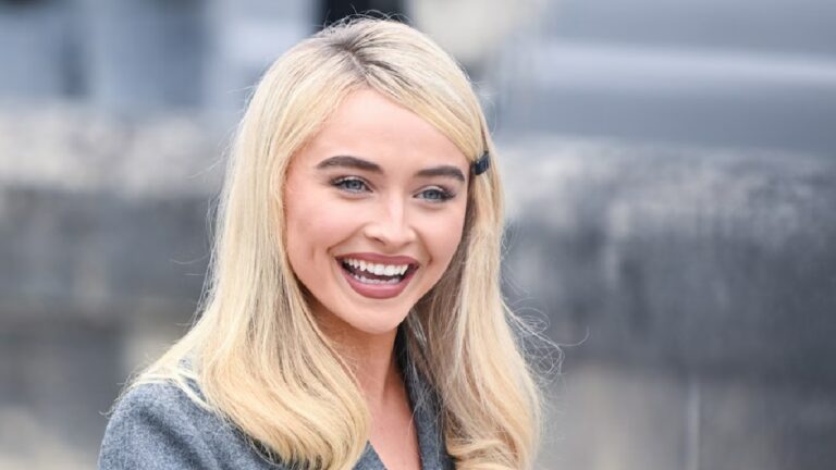 Sabrina Carpenter sorri amplamente em ambiente externo, com cabelo solto preso por um grampo e vestindo roupa cinza, transmitindo simpatia e alegria