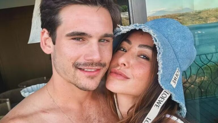 A imagem mostra um casal sorridente em um momento descontraído. A mulher usa um chapéu de tecido jeans e está próxima ao homem, que aparece sem camisa. O cenário ao fundo sugere um ambiente litorâneo ou de férias, transmitindo uma atmosfera de relaxamento e carinho