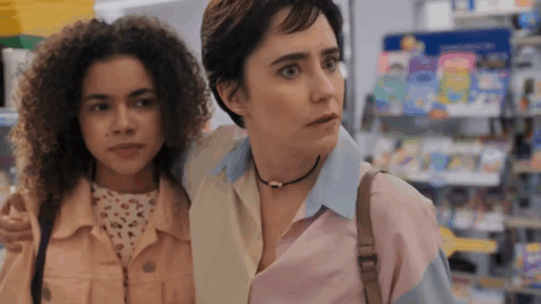 Mulher de expressão surpresa abraça uma jovem de jaqueta rosa em ambiente interno bem iluminado, com prateleiras de produtos coloridos ao fundo