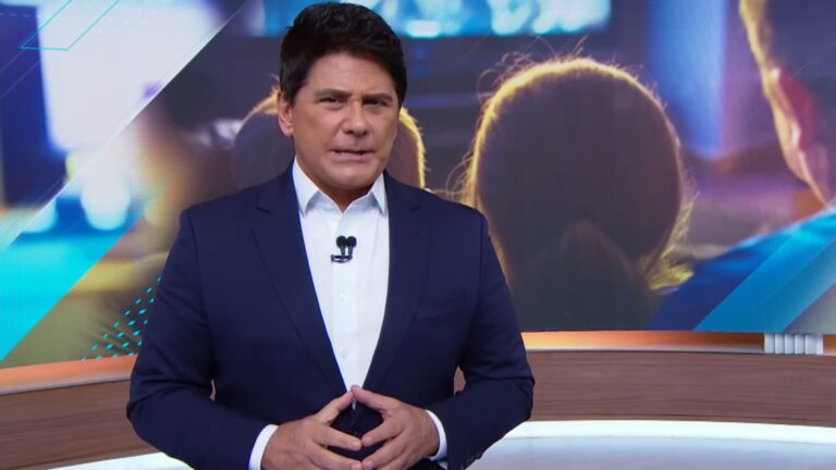 A imagem mostra César Filho no estúdio do SBT Brasil. Ele está vestindo um terno azul escuro com camisa branca e segura as mãos à frente do corpo enquanto fala. Atrás dele, há um grande telão com uma imagem desfocada de pessoas assistindo uma TV, em tons de azul e dourado. O cenário é moderno e bem iluminado, típico de programas jornalísticos