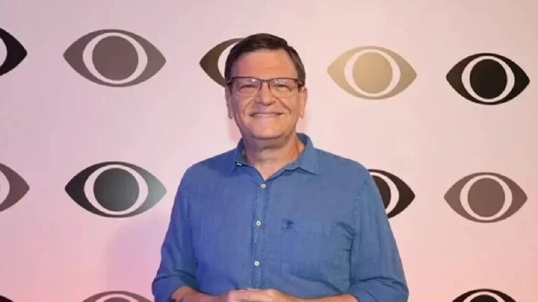 Homem sorri para a câmera diante de um painel com o logotipo repetido de uma emissora de televisão, vestindo camisa azul e óculos, em ambiente de evento ou estúdio