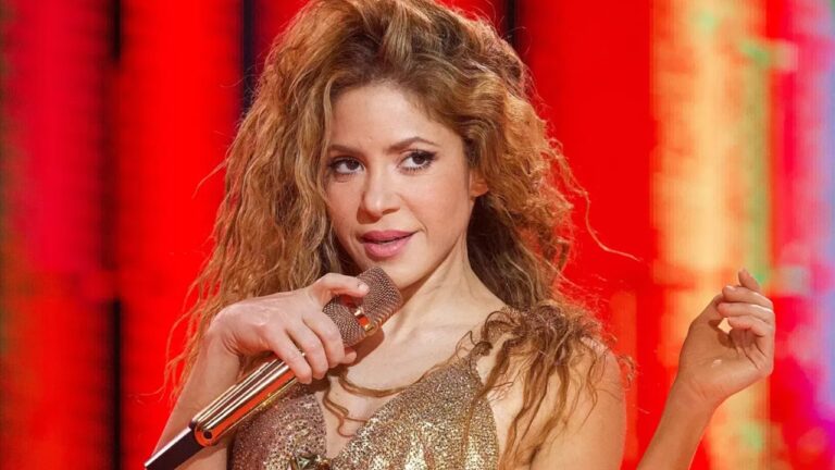 A imagem mostra uma mulher cantando em um palco iluminado por luzes intensas em tons de vermelho e laranja. Ela segura um microfone metálico dourado próximo ao rosto e usa um vestido brilhante na mesma paleta dourada. Seu cabelo é longo, volumoso e cacheado, e ela mantém uma expressão confiante enquanto performa