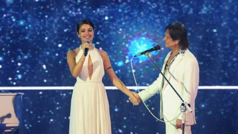 Cantora de vestido branco e cantor de terno claro se apresentam juntos no palco, de mãos dadas, diante de fundo iluminado com luzes azuis que lembram o céu estrelado