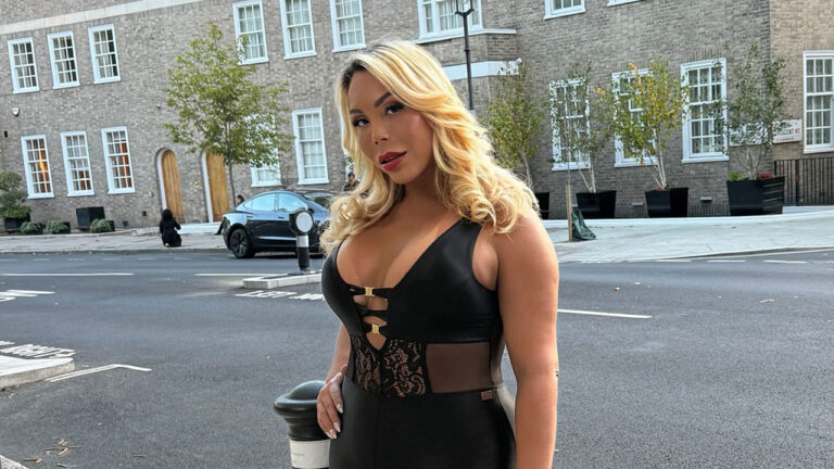 Suellen Carey posando na rua, com cabelo loiro, maquiagem forte e vestindo um body preto de couro e renda