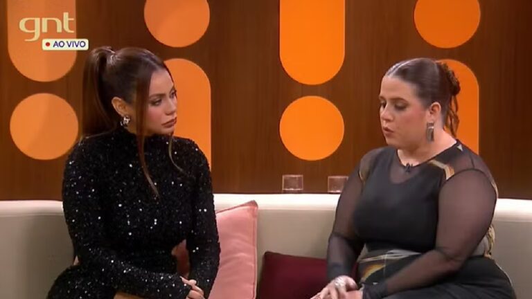 Cena de um programa do canal GNT exibido ao vivo mostra duas mulheres sentadas em um sofá conversando seriamente em um cenário com fundo laranja e almofadas coloridas