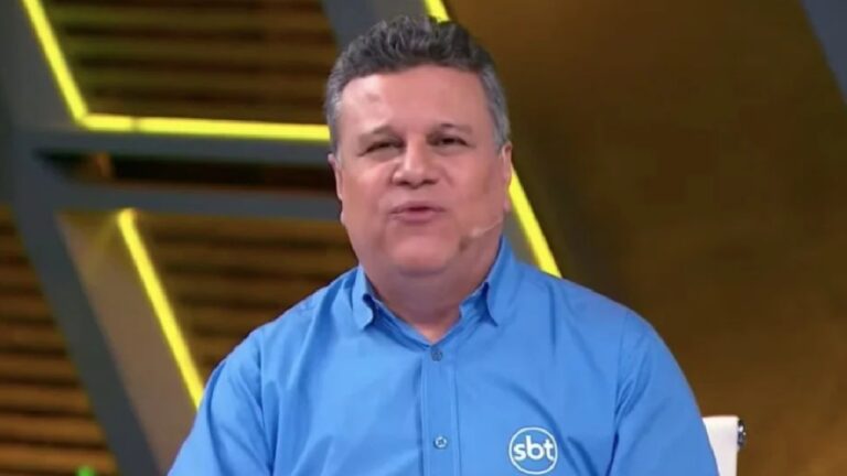 Homem de camisa azul com o logotipo do SBT fala em estúdio iluminado, usando microfone de orelha e posicionado diante de cenário com linhas amarelas