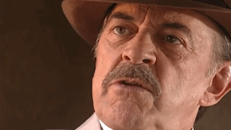 Close-up de um homem mais velho usando chapéu marrom e terno claro, com expressão séria e olhar fixo. A iluminação suave destaca os traços de seu rosto, transmitindo intensidade e emoção à cena