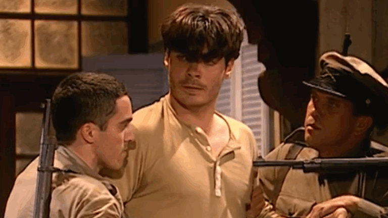 A imagem mostra uma cena de época ambientada em um local interno, com iluminação amarelada. No centro, um homem de expressão séria e cabelos despenteados é segurado por dois soldados uniformizados e armados com fuzis, que o mantêm sob vigilância. O clima é tenso, sugerindo uma prisão ou confronto. Ao fundo, é possível ver uma janela e venezianas parcialmente abertas