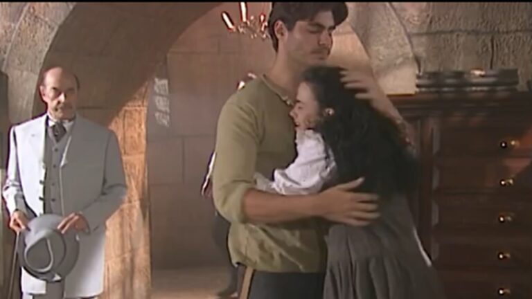 A imagem mostra uma cena de época ambientada em um local de pedra, provavelmente uma prisão ou casa antiga. Um homem de camisa clara abraça uma mulher de cabelos longos e escuros, que parece emocionada. Ao fundo, outro homem de terno claro e chapéu observa a cena com expressão séria