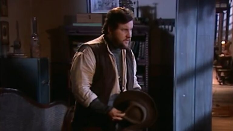 Homem barbado com chapéu nas mãos em sala rústica iluminada pela porta aberta em cena de época