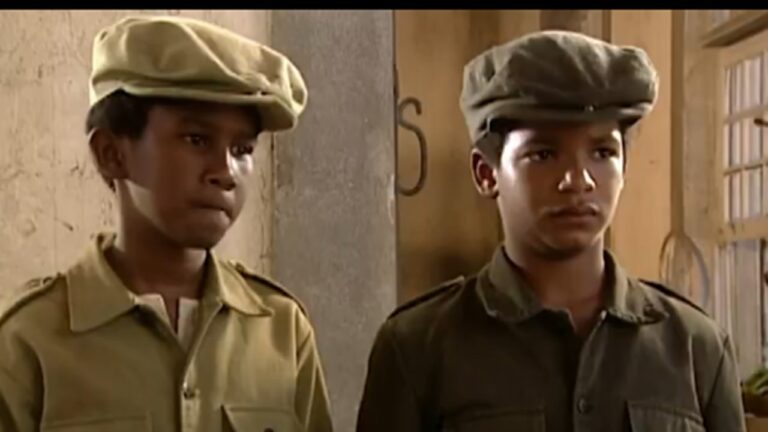 Dois meninos com boinas e camisas de época observam atentos em ambiente simples de madeira