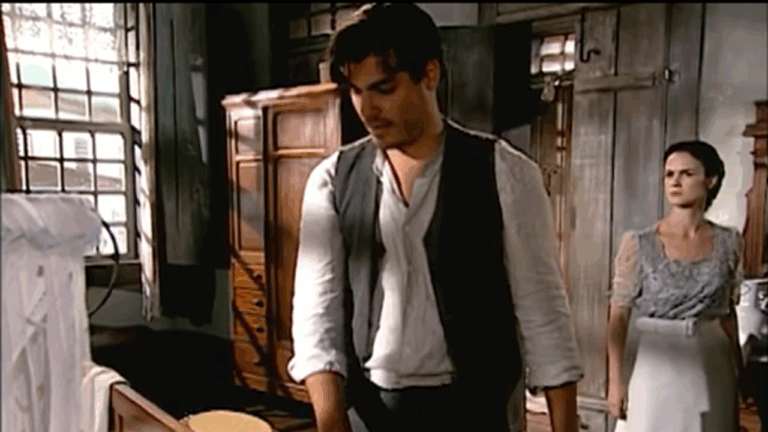 Cena de época mostra um homem e uma mulher em um quarto simples, iluminado pela luz natural que entra pelas janelas de madeira. O homem está cabisbaixo, vestindo camisa clara e colete escuro, enquanto a mulher, com expressão séria, observa ao fundo, usando um vestido antigo com avental branco