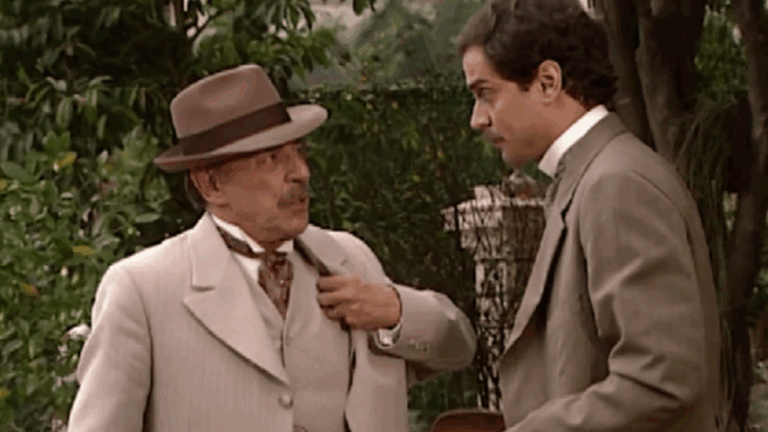 Dois homens vestidos com ternos de época conversam em um ambiente externo com vegetação ao fundo. Um deles, mais velho e usando chapéu, gesticula enquanto fala; o outro, mais jovem, escuta atentamente. A cena transmite um ar histórico e formal, típico de produções de época