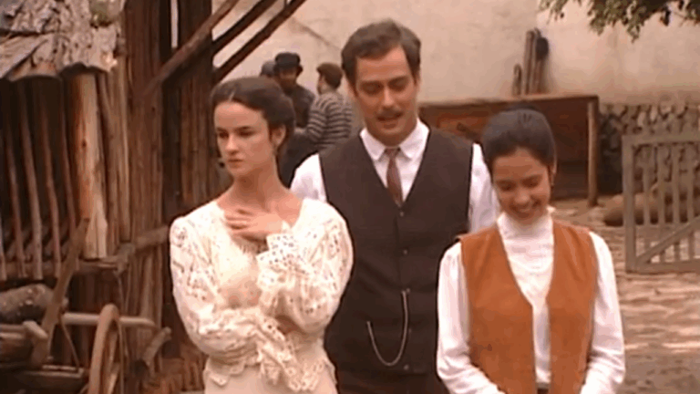 A imagem mostra três pessoas em um ambiente rural, com construções de madeira e uma carroça ao fundo. As roupas dos personagens remetem ao início do século XX: o homem veste colete e gravata, enquanto as mulheres usam vestidos e blusas de renda, com penteados presos. A cena parece fazer parte de uma produção de época, transmitindo um clima de drama e romance histórico