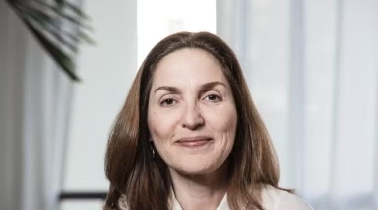 A imagem mostra uma mulher de cabelos castanhos e lisos, sorrindo levemente. Ela está em um ambiente interno com cortinas claras e iluminação suave, transmitindo uma sensação profissional e acolhedora