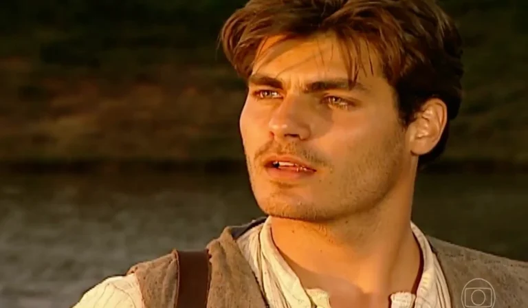 Homem jovem com cabelos castanhos e barba rala aparece em close sob a luz dourada do pôr do sol. Ele olha para o horizonte com expressão séria. Veste camisa clara de linho e colete rústico, com uma alça de mochila sobre o ombro. Ao fundo, vê-se um rio desfocado e vegetação escura