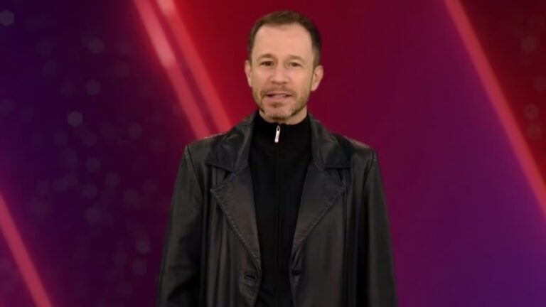 A imagem mostra Tiago Leifert em pé diante de um fundo com tons de vermelho e roxo. Ele veste uma jaqueta de couro preta sobre uma blusa escura com zíper. Ele está no estúdio do programa The Voice Brasil, exibido pelo SBT