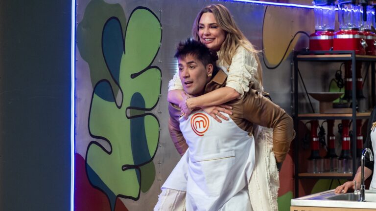 Foto de uma cena descontraída no estúdio do MasterChef Celebridades: o cantor Tiago Piquilo, vestido com um avental do MasterChef, carrega a jornalista Rachel Sheherazade nas costas, ambos demonstrando alegria. O ambiente colorido e iluminado reforça o clima leve e divertido do momento