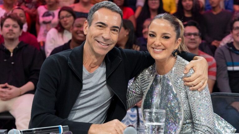 A imagem mostra duas pessoas sorridentes sentadas lado a lado em um estúdio, com o homem abraçando a mulher. Ambos parecem felizes e estão bem vestidos — ela usa uma roupa brilhante prateada, e ele veste um casaco escuro com camiseta cinza. Ao fundo, há uma plateia observando, sugerindo que a cena ocorre durante a gravação de um programa de TV