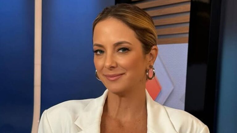 Apresentadora de blazer branco sorri em estúdio jornalístico com fundo azul e elementos geométricos iluminados