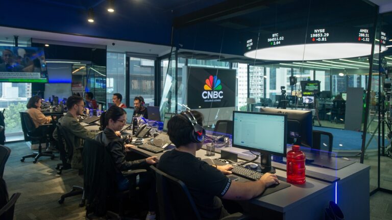 A imagem mostra uma redação moderna da CNBC, com várias pessoas trabalhando em frente a computadores. O ambiente tem paredes de vidro, vista para prédios da cidade e uma grande tela com o logotipo da emissora