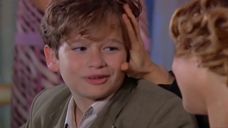 A imagem mostra um garoto de cabelos castanhos claros sendo acariciado na cabeça por uma mulher. Ele está sorrindo e usa um paletó verde-oliva com camisa clara por baixo. A cena parece ser de uma novela ou série, com um ambiente interno e cores suaves ao fundo