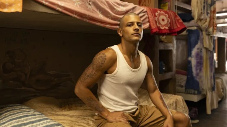 A imagem mostra um homem de cabeça raspada e tatuagens nos braços, sentado em uma cama simples dentro de um quarto coletivo com beliches. Ele veste uma regata branca e calça bege, olhando fixamente para a frente com expressão séria
