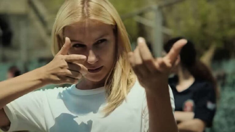 A imagem mostra uma mulher loira usando uma camiseta branca, fazendo um gesto com as mãos como se estivesse mirando com uma arma imaginária. Ela parece concentrada, e o cenário é externo, com pessoas e árvores desfocadas ao fundo, sugerindo uma cena de um filme ou série