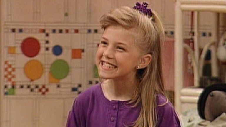 A imagem mostra uma menina sorridente com cabelo loiro preso por um elástico roxo e usando uma blusa da mesma cor. O cenário ao fundo parece ser um quarto infantil, com parede decorada com figuras geométricas coloridas e móveis claros, remetendo a um ambiente de série de TV familiar dos anos 1990