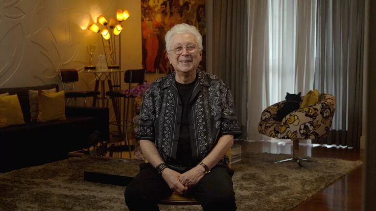 A imagem mostra um homem mais velho sentado em uma sala de estar aconchegante, sorrindo para a câmera. Ele usa óculos redondos, uma camisa estampada preta e calça escura. O ambiente ao redor é elegante e bem decorado: há um tapete felpudo, uma poltrona estampada onde um gato preto está deitado, cortinas longas e uma parede com relevo decorativo. No fundo, luminárias amarelas e objetos de decoração dourados complementam a cena, criando uma atmosfera acolhedora e refinada