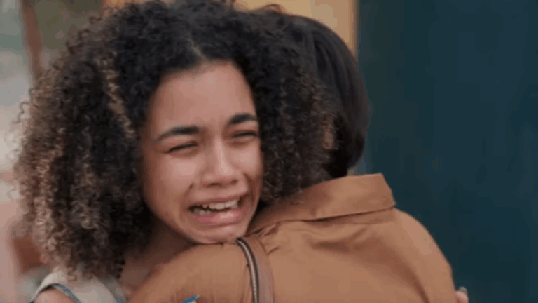 A imagem mostra uma jovem de cabelos cacheados chorando intensamente enquanto abraça outra pessoa, que veste uma camisa marrom. A expressão da jovem transmite dor e emoção, sugerindo um momento de despedida ou reencontro comovente