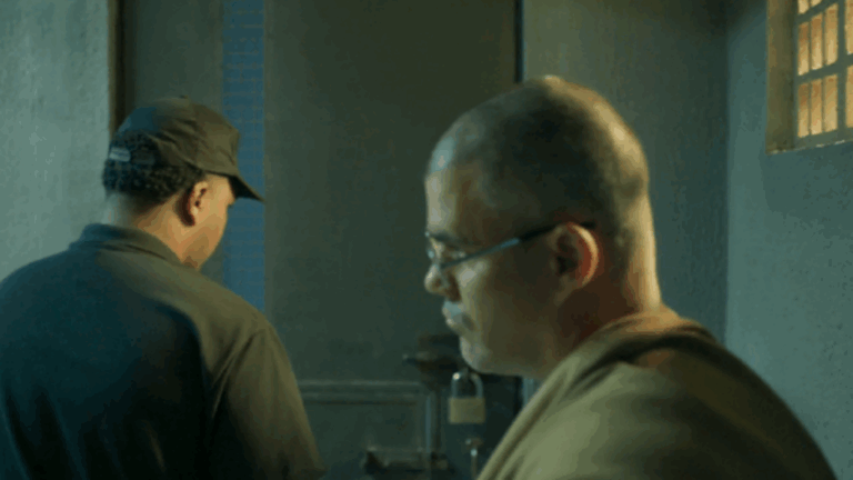 Cena mostra dois homens em um ambiente com iluminação fria e paredes de concreto, sugerindo um local como uma prisão ou uma sala de vigilância. Um deles está de boné, de costas, enquanto o outro, de óculos e expressão séria, parece refletir ou conversar sobre algo importante