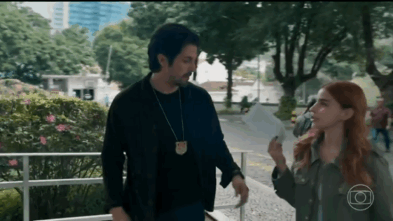 A imagem mostra uma cena ao ar livre, com um homem e uma mulher caminhando lado a lado em um local arborizado. O homem usa um crachá pendurado no pescoço, sugerindo ser um policial ou investigador, enquanto a mulher segura um papel e parece estar explicando algo. O clima é de conversa séria e investigação